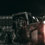 A black Labrador retriever lies in front of a vehicle with the GUNNER G1™ KENNEL - BLACK, a tough metal safe dog crate, illuminated by warm light and surrounded by darkness and a slight haze.